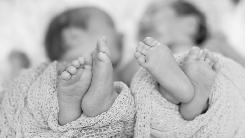 Two newborn twin babies lying side by side, one with eyes open looking at the camera, the other sleeping peacefully. Both wrapped in soft white blankets.